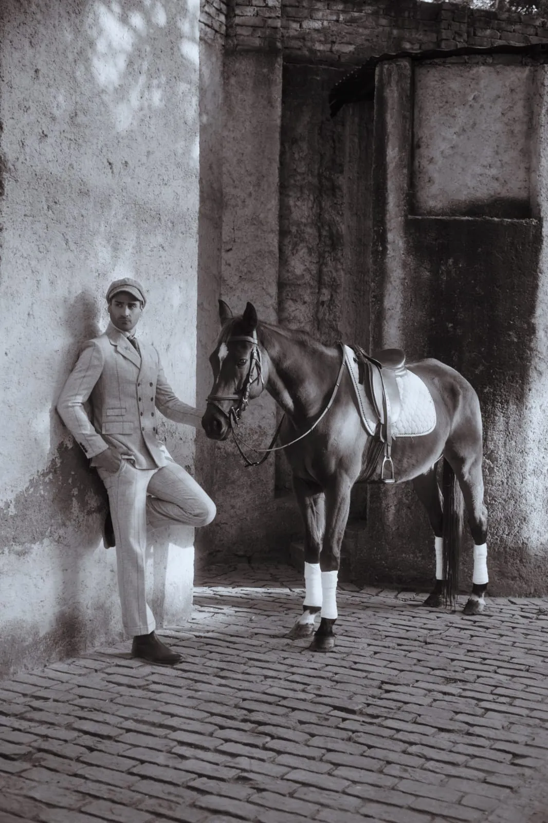 Sepia portrait of a rider in tweed beside a dark horse, weathered stable wall behind.
