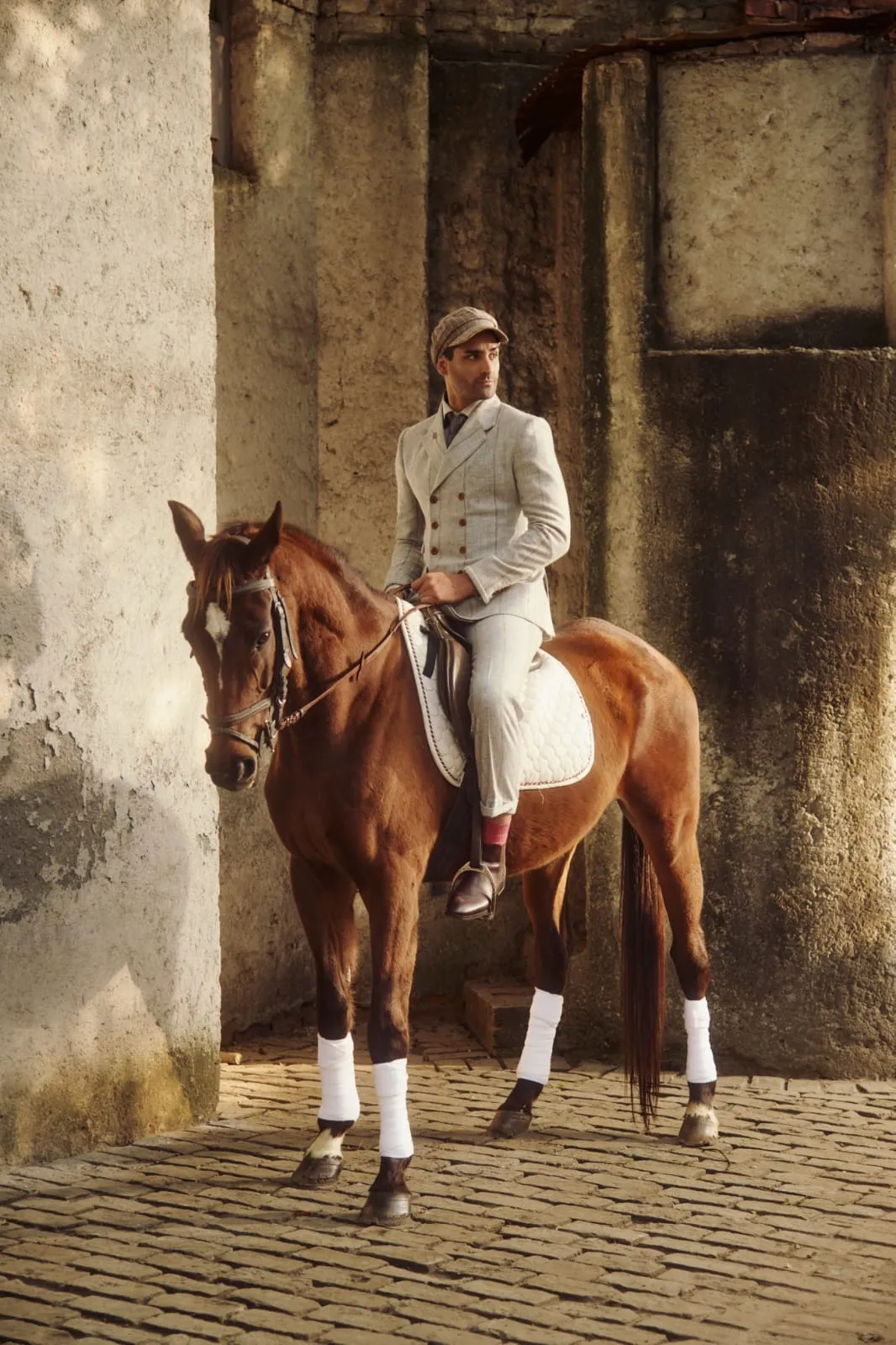 A rider in a tweed three-piece suit and flat cap, mounted on a chestnut horse on cobblestone.