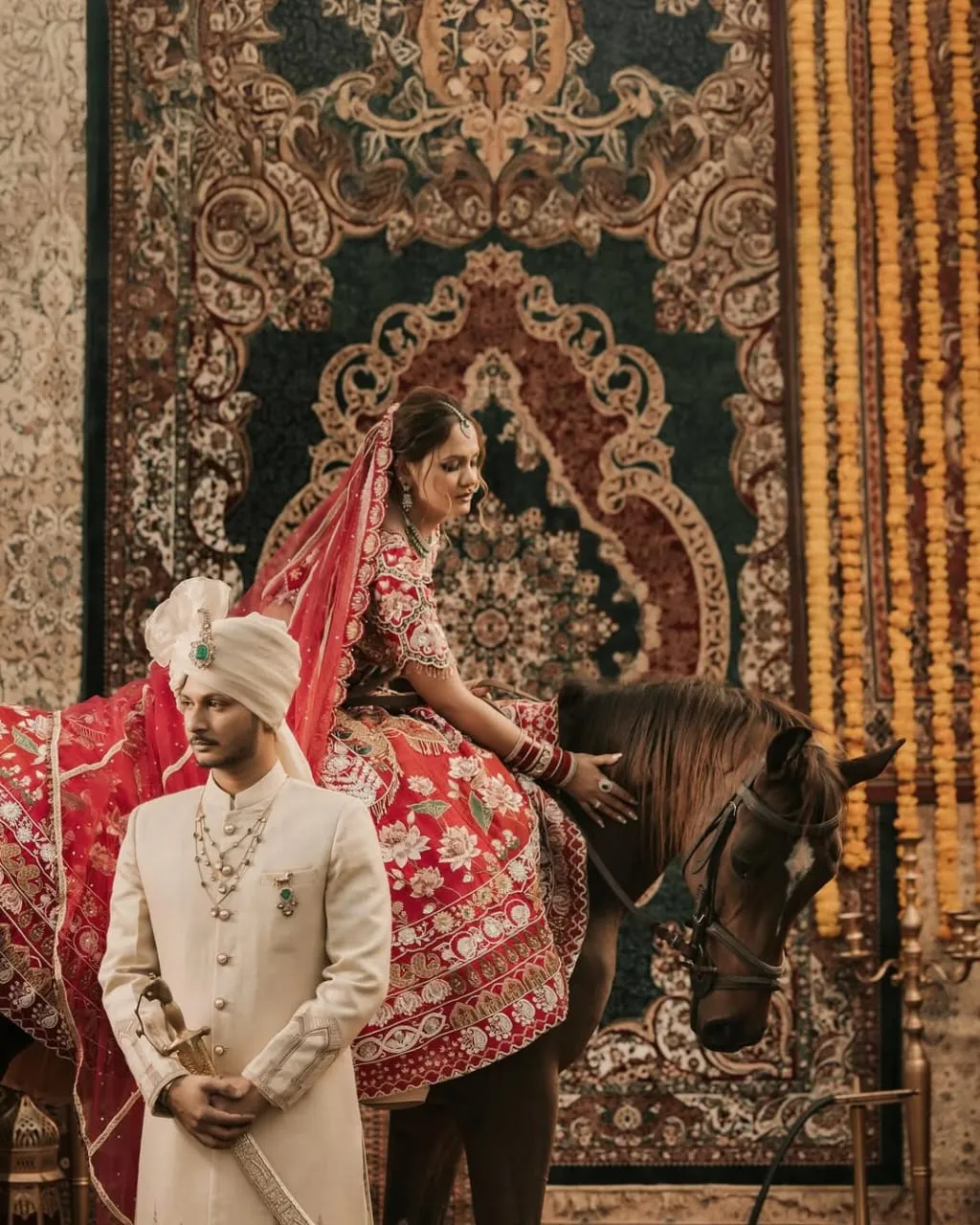 A bride in red bridal lehenga sidesaddle on a dark horse, framed by a richly patterned tapestry and brass diyas during a pre-wedding shoot at FHRC.