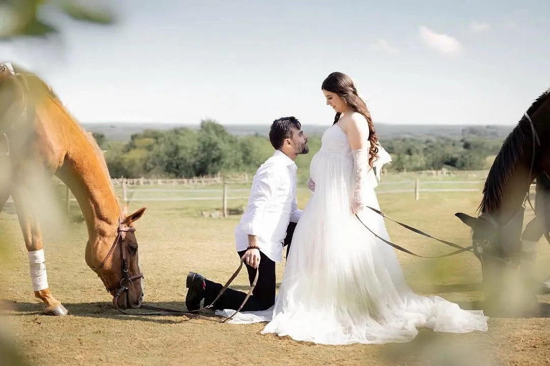 A couple in a flowing white dress, kneeling and standing beside grazing horses in an open meadow.