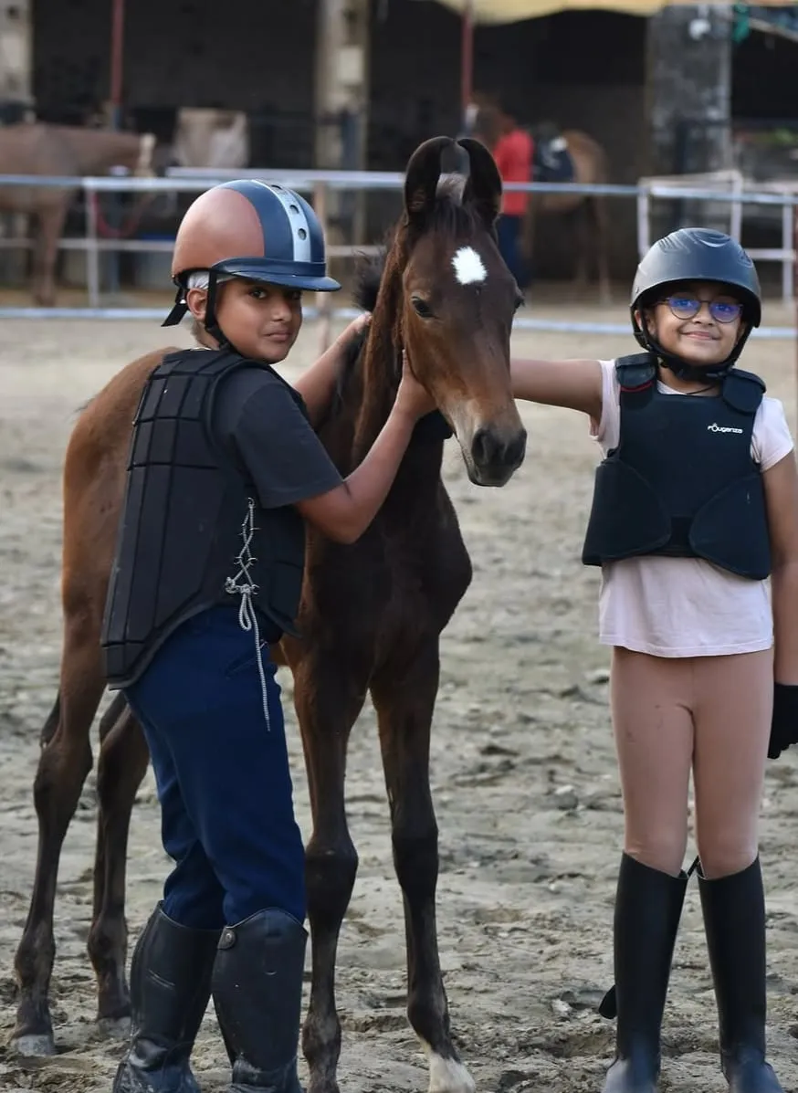 Two young beginner riders in helmets and body protectors stand beside a foal at FHRC, learning safe handling.